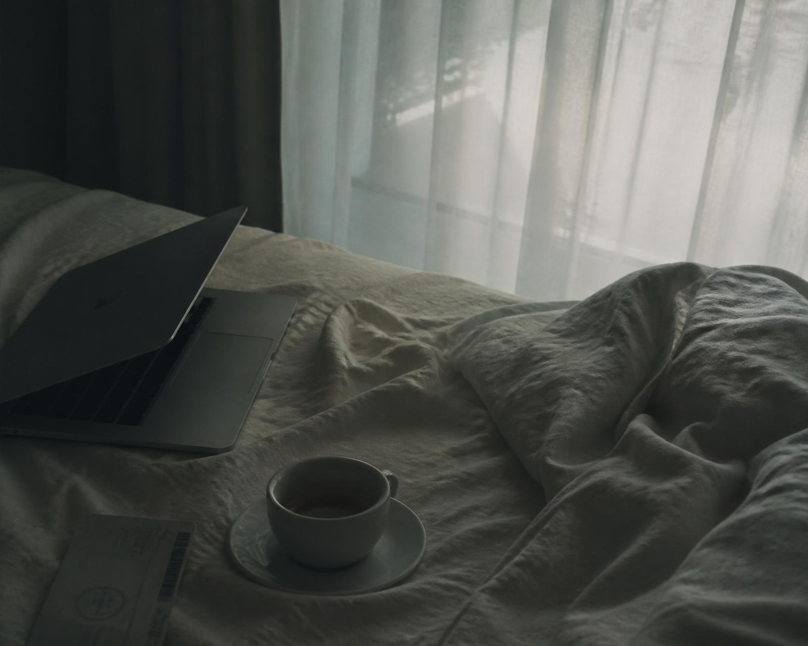 A rumpled linen bed with a half-open laptop, an espresso cup, and a boarding pass in cool dawn light.